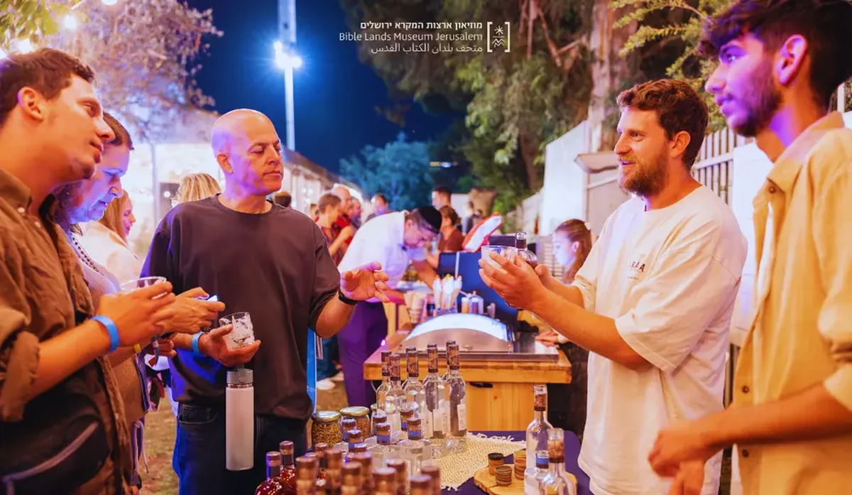 אנשים עומדים ליד דוכן משקאות בפסטיבל אלכוהול, עם בקבוקי זכוכית שקופים וצבעוניים