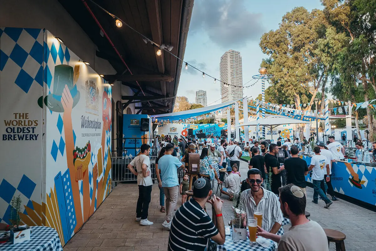 פסטיבל בירה חוצות עם אנשים רבים, דוכני בירה, וקישוטים בכחול-לבן.